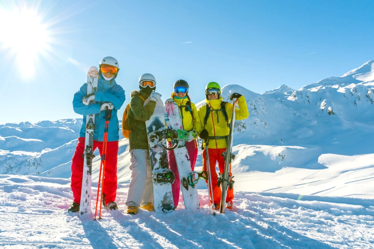 Four happy friends snowboarders and skiers are having fun on a ski slope with ski and snowboards in a sunny day and mountains view background. how to budget for a ski vacation ski group rentals